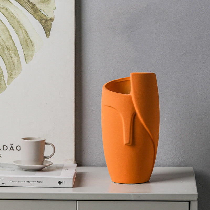 Orange vase with a unique design on a surface next to a cup and books, with a gray wall and framed artwork in the background.