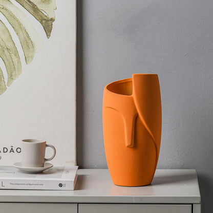 Orange vase with a unique design on a surface next to a cup and books, with a gray wall and framed artwork in the background.