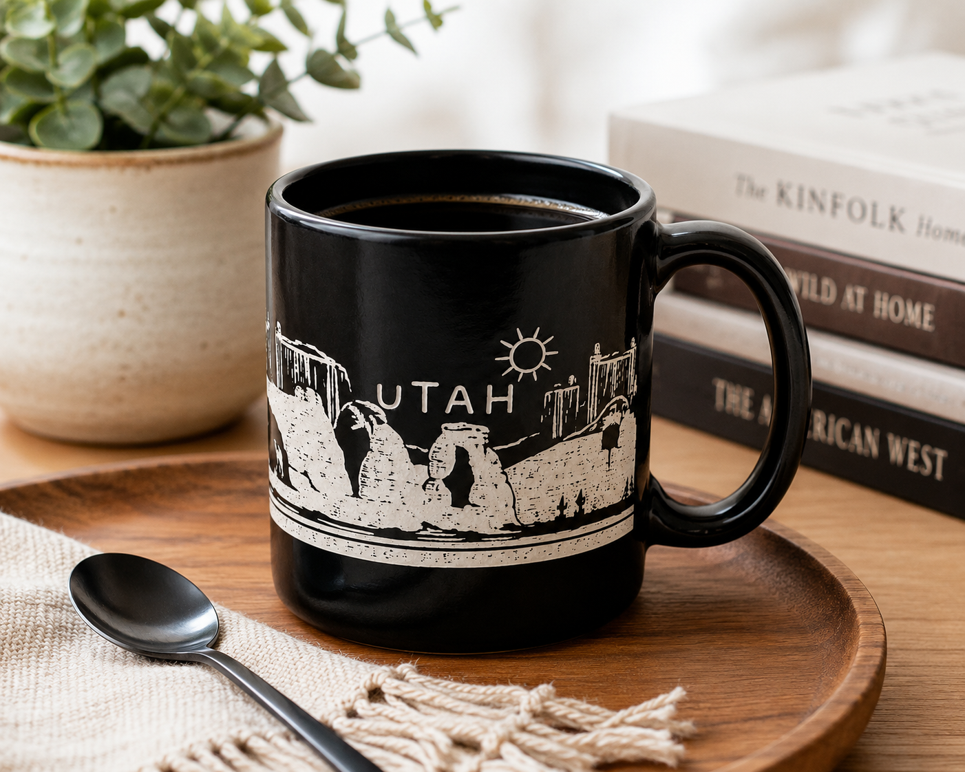 A black ceramic mug with a white line-art graphic of Utah landmarks and the word 'UTAH' on the side, styled on a wooden tray with a spoon, books, and a potted plant in the background.