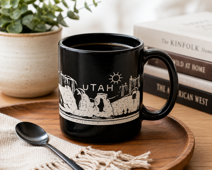 A black ceramic mug with a white line-art graphic of Utah landmarks and the word 'UTAH' on the side, styled on a wooden tray with a spoon, books, and a potted plant in the background.