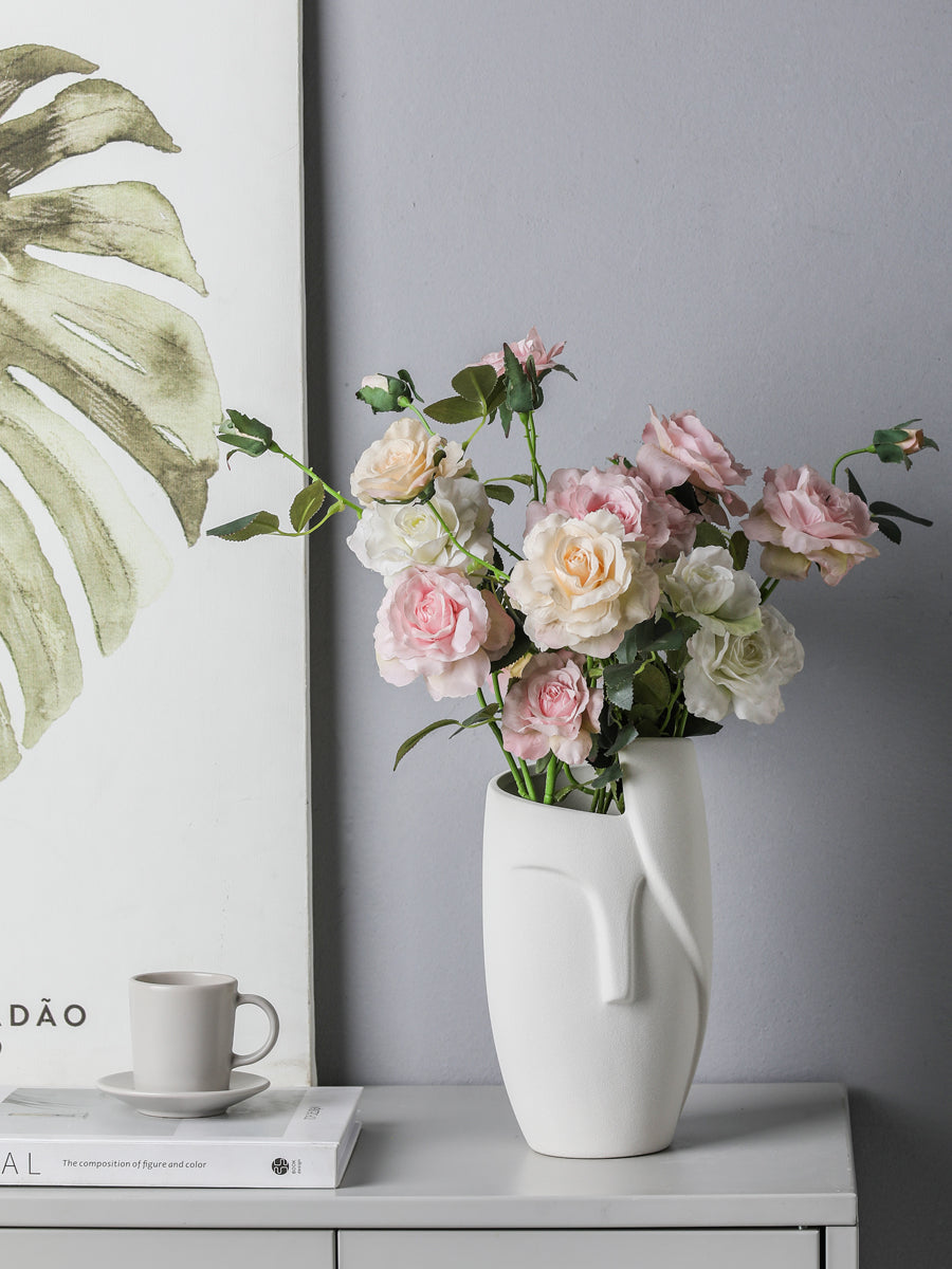 White vase with pink flowers on a gray surface next to a framed plant print.
