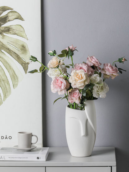 White vase with pink flowers on a gray surface next to a framed plant print.
