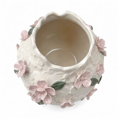 An off-white textured ceramic vase decorated with 3D light pink flowers and green leaves, shown from a top-down angle on a white background.