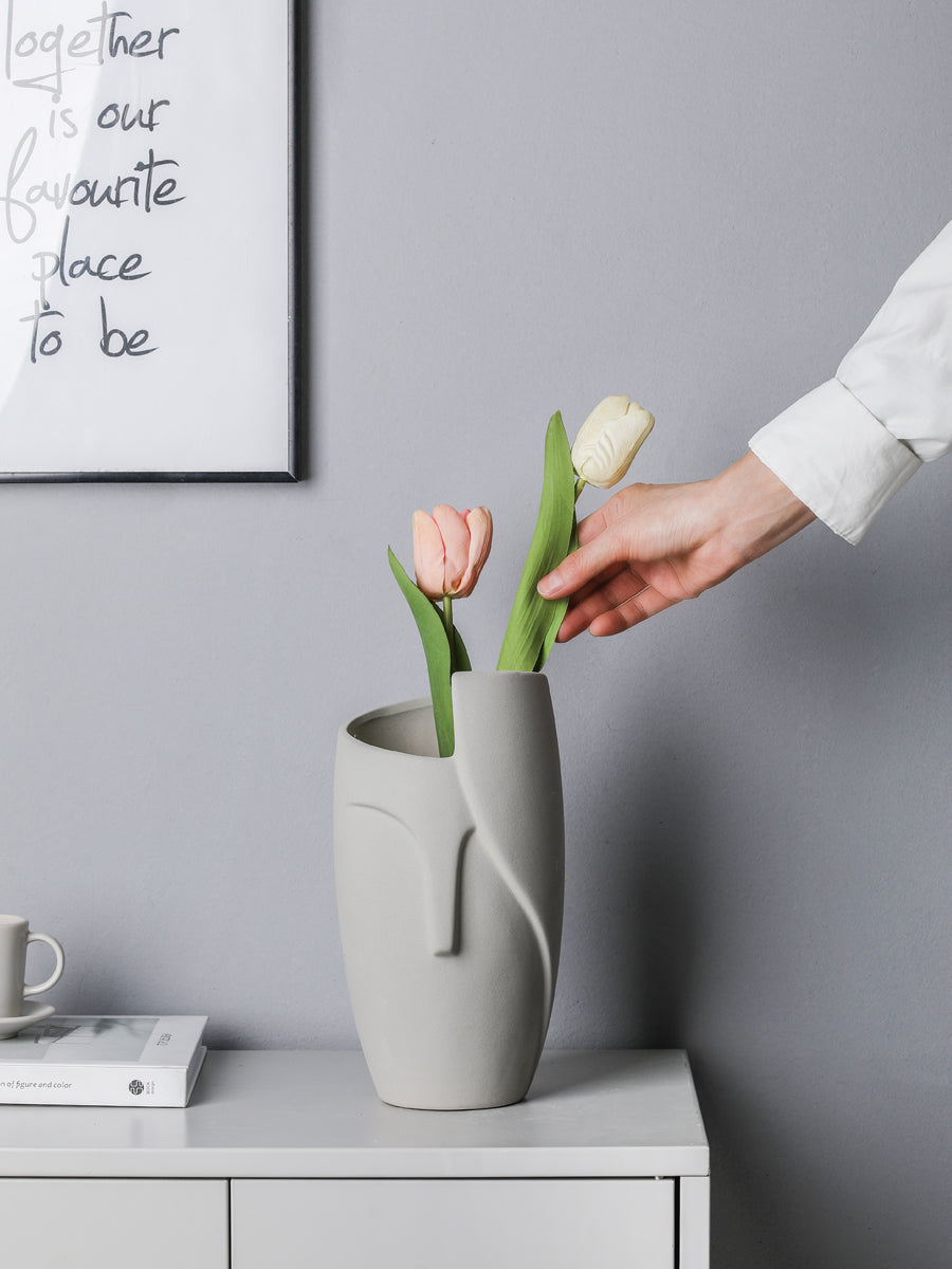 Person arranging tulips in a gray vase on a table with a gray wall and framed text in the background.