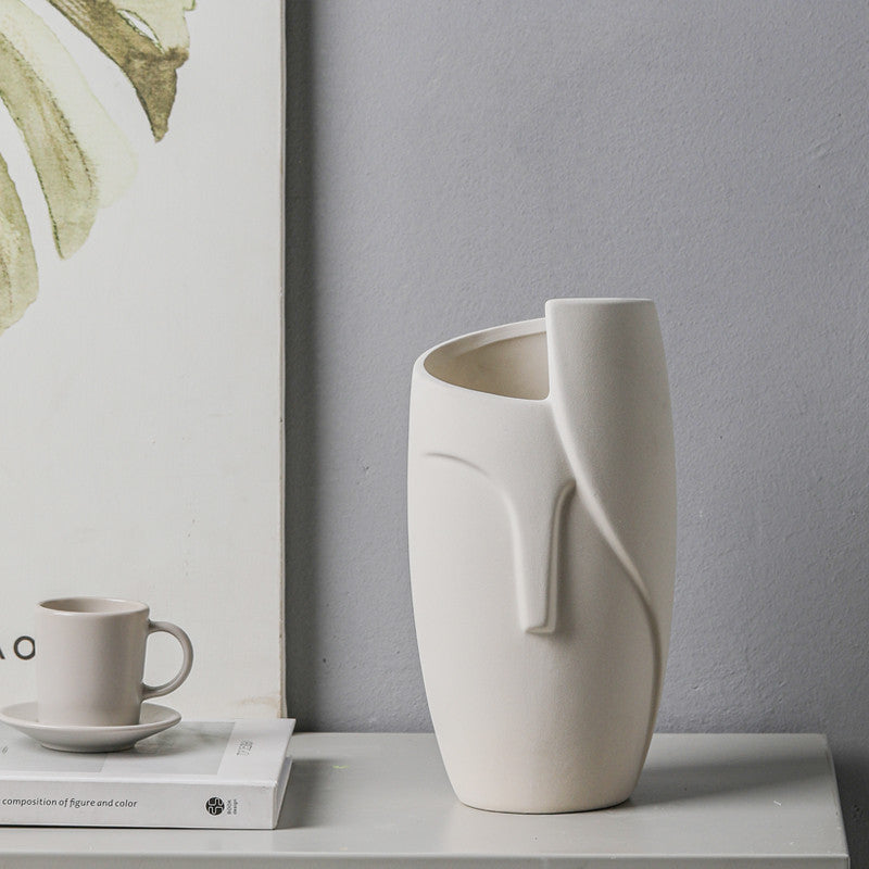 White ceramic vase on a table with a gray wall and framed artwork in the background