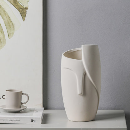 White ceramic vase on a table with a gray wall and framed artwork in the background