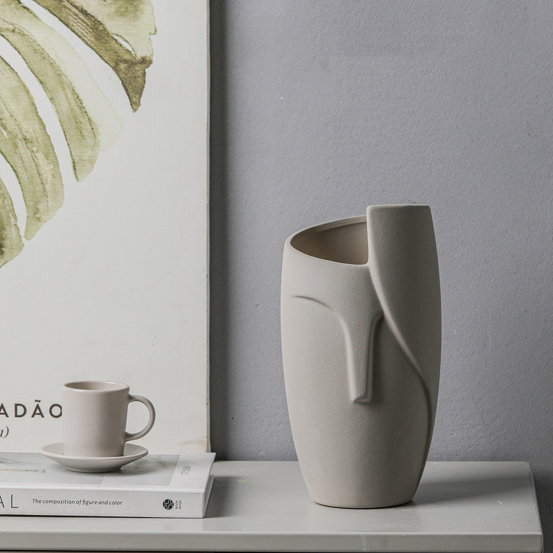 Gray face-shaped vase on a surface with a cup and books in the background